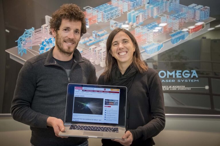 Photo of two people posing in front of a poster while holding a laptop computer that is displaying a journal article.