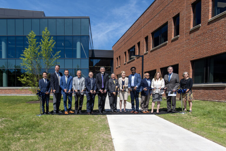 Group of people in business attire standing outdoors in front of a brick and glass building during a ribbon-cutting ceremony.