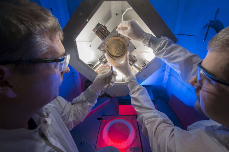 Photo of two people in lab attire handling a piece of lab equipment.