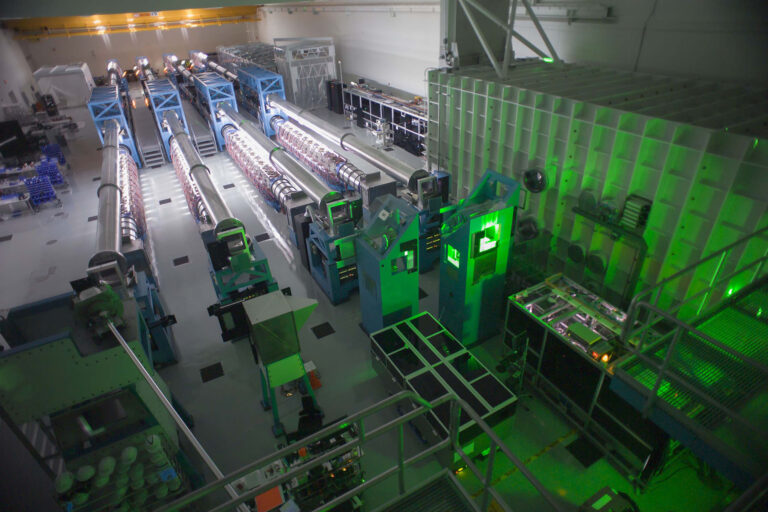 Large laser facility hall with rows of high-energy laser beamlines glowing green and complex equipment arranged along elevated walkways.