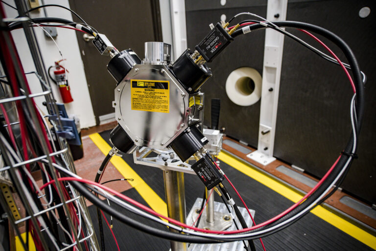 Close-up of a diagnostic instrument with multiple fiber-optic probes arranged around a circular target area, mounted on a lab platform with cables running outward.