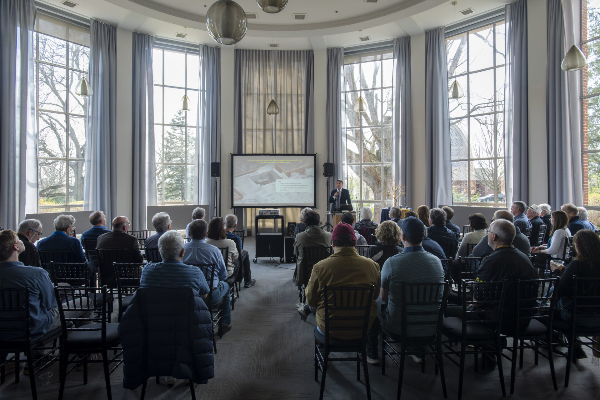 Audience seated in rows of chairs attending a presentation in a large, circular event space with floor-to-ceiling windows and tall gray curtains. A speaker stands at a podium in front of a projection screen, while natural light fills the room through the expansive windows overlooking trees outside. Modern pendant lights hang from the high ceiling above the gathering.