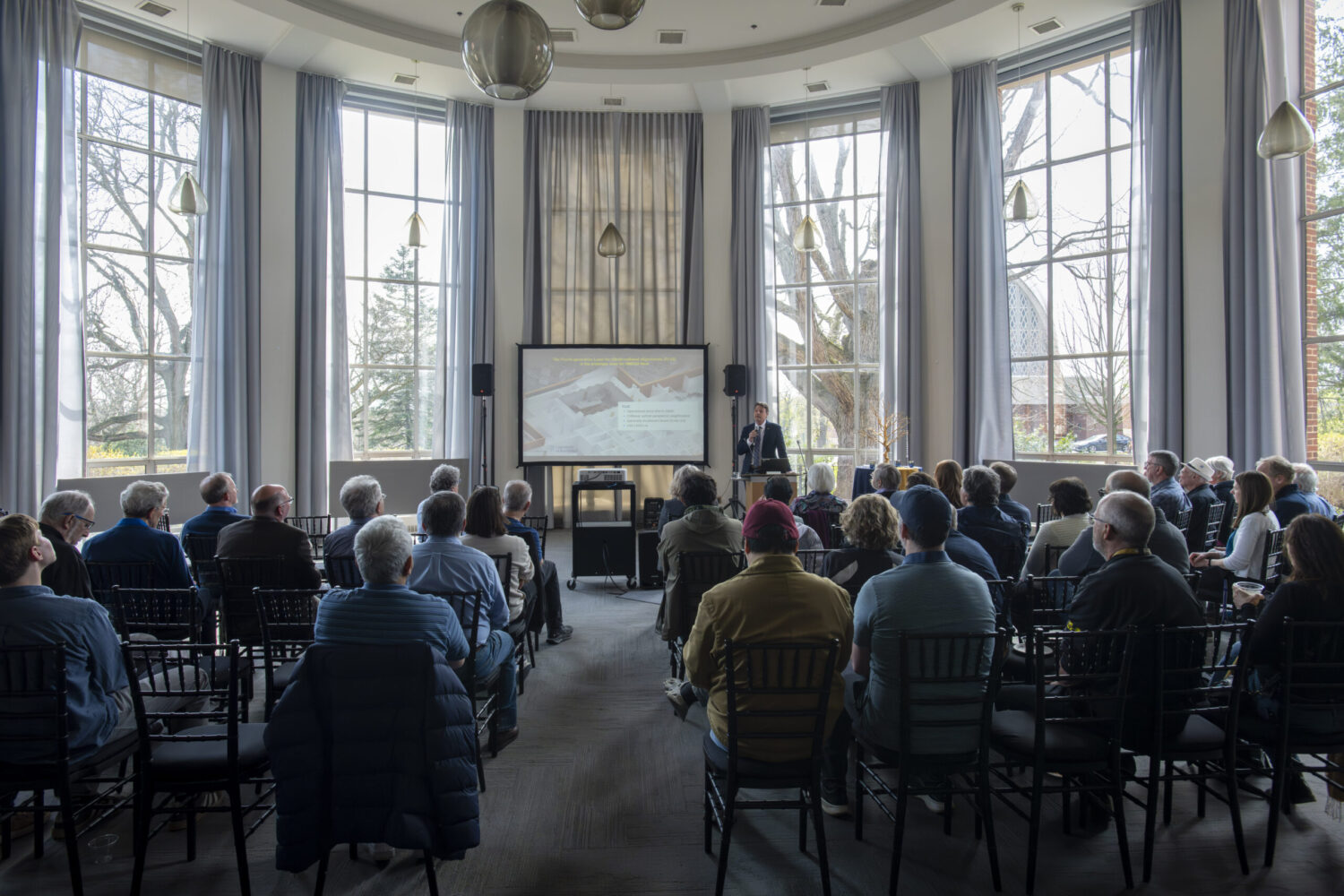 Audience seated in rows of chairs attending a presentation in a large, circular event space with floor-to-ceiling windows and tall gray curtains. A speaker stands at a podium in front of a projection screen, while natural light fills the room through the expansive windows overlooking trees outside. Modern pendant lights hang from the high ceiling above the gathering.